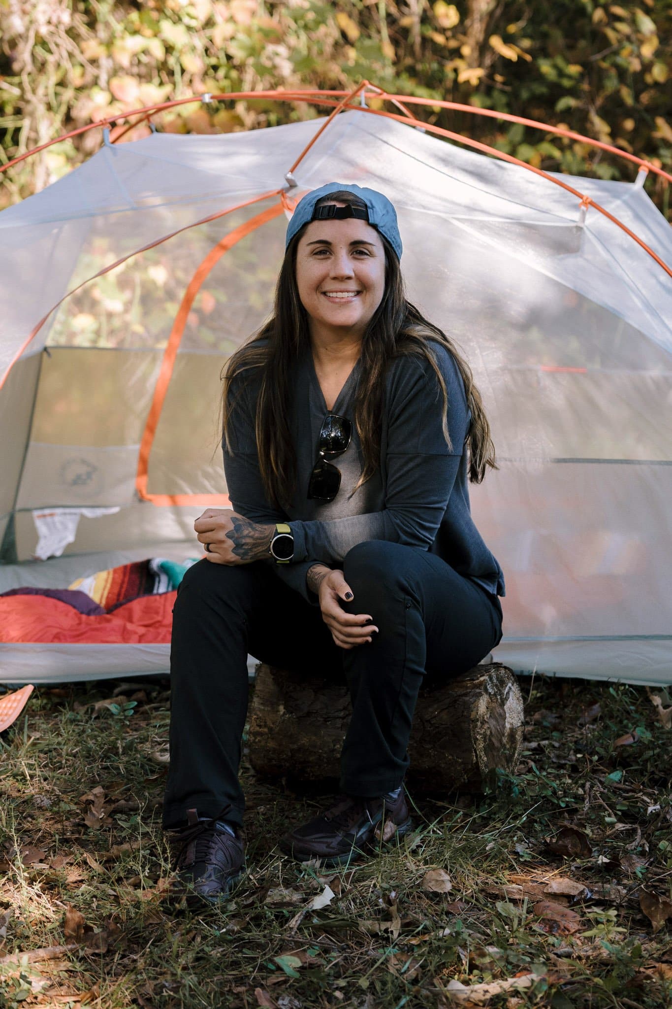 Bethany, the author of the post Camping Progressions to Get You Started, is sitting on a log in front of her tent smiling. 